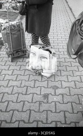 Un sac arborant une photographie de portrait se tient à côté des pieds d'une femme dans une scène de rue analogique en noir et blanc à contraste élevé. Banque D'Images