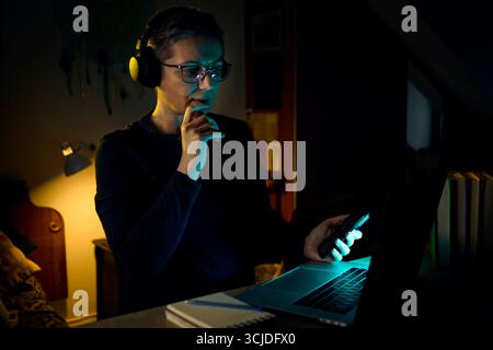 Une femme s'assoit dans son bureau à domicile la nuit avec des écouteurs, se concentrant sur l'écran de son ordinateur portable. La scène reflète le travail à distance, les outils de communication modernes Banque D'Images