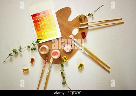 Vue de dessus de la palette en bois d’un artiste avec des peintures à l’aquarelle, des pinceaux et du papier échantillon dans des tons chauds. Espace de travail créatif pose plate pour la peinture, art. Banque D'Images