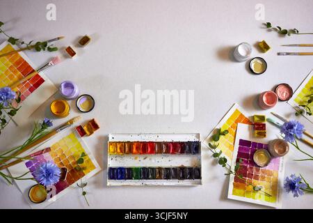 Pose à plat de palette d'aquarelles colorées, de pinceaux, de pots de peinture, d'éléments floraux et d'échantillons peints sur papier. Fond d'espace de travail artistique, perfec Banque D'Images