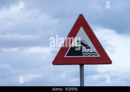 Panneau d'avertissement triangulaire rouge indiquant le danger de la voiture tomber dans l'eau, posé contre le ciel nuageux Banque D'Images
