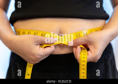 Une femme vérifiant sa taille avec un ruban à mesurer, symbolisant la santé, la forme physique, un régime, la graisse corporelle, et les objectifs de perte de poids Banque D'Images