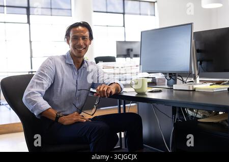 Homme d'affaires confiant assis au bureau dans un bureau moderne, tenant des lunettes, souriant Banque D'Images