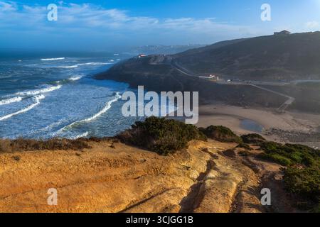 Une vue imprenable sur la côte avec des falaises dorées, des vagues turquoises qui s'écrasent sur une plage de sable et une route sinueuse le long du rivage sous un SK bleu vif Banque D'Images