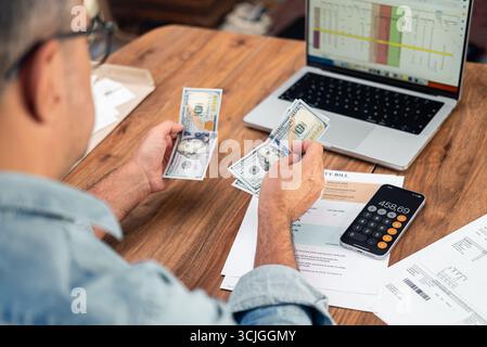Homme mature avec ordinateur portable et factures de services publics compte des billets de dollar pour les paiements assis à table dans le bureau Banque D'Images