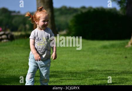 Adorable jeune fille aux cheveux rouges avec des queues de cheval souriantes et jouant sur l'herbe verte dans le jardin d'été ensoleillé. Joie de l'enfance et style de vie en plein air. Banque D'Images