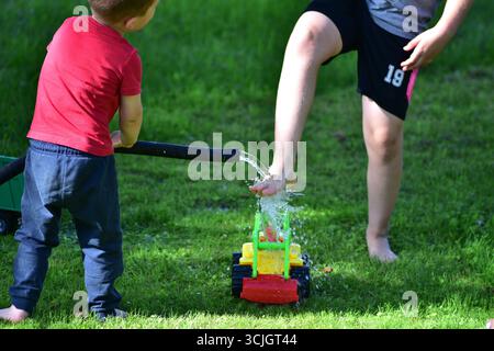 Enfants jouant avec le tuyau d'arrosage et le tracteur jouet, éclaboussant de l'eau sur l'herbe en été. Activités d'enfance en plein air amusantes et loisirs en famille. Banque D'Images