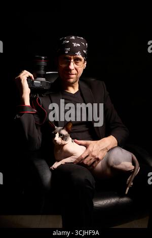 Un photographe masculin portant un bandana pose dans un studio sombre tout en tenant un chat Sphynx canadien et un appareil photo. Le portrait dramatique met en valeur la créativité Banque D'Images