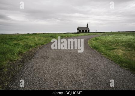 Église en bois noir Budakirkja se dresse dans un paysage rural avec une route de campagne menant à travers des champs verts sous un ciel nuageux dans le Village Budh Banque D'Images