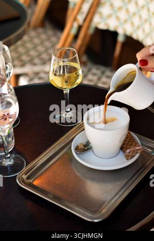 Plan vertical d'une main de femme versant du café dans de la mousse de lait, avec du vin blanc sur la table, Paris vibe de chaises tissées. Idéal pour les visuels de voyage, les scènes culinaires ou le style de vie chic avec un attrait confortable. Banque D'Images