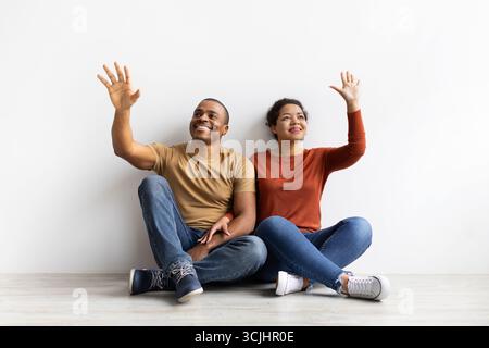Couple afro-américain souriant assis sur le sol, levant les mains rêveur, imaginant quelque chose Banque D'Images