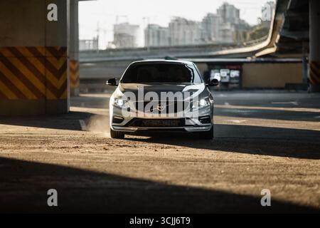 volvo S60 argenté sous un pont au coucher du soleil. Vue avant de la berline sportive suédoise sous les rayons du soleil couchant, rangée de piliers en béton sur la droite Banque D'Images