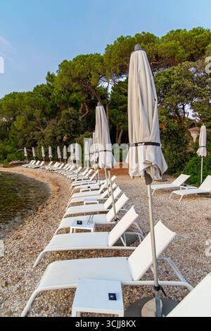 Chaises longues vides et parasols fermés sur la plage de galets. Une rangée de chaises longues blanches avec parasols fermés sur une plage de galets tranquille bordée d'arbres Banque D'Images