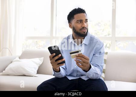 Pensif jeune homme prenant la décision de paiement avec téléphone portable de carte bancaire Banque D'Images