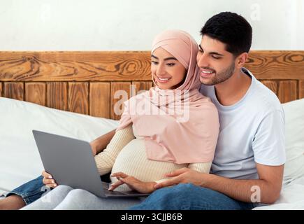 Joyeux couple arabe enceinte utilisant un ordinateur portable, regardant le film ensemble tout en se reposant sur le lit Banque D'Images