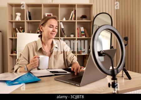 Une femme travaillant sur un ordinateur portable, tenant une tasse à café, assise dans un espace de travail bien organisé avec des étagères et des fournitures de bureau. Une lueur d'anneau Banque D'Images
