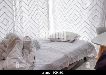 Lit confortable avec literie blanche grondée et oreillers moelleux, baigné de lumière naturelle par les rideaux à motifs, évoquant une ambiance reposante et relaxante Banque D'Images