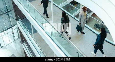 Un groupe de gens d'affaires divers marchant vigoureusement dans un espace de bureau contemporain avec des balustrades en verre et une architecture élégante. L'image capture le Banque D'Images