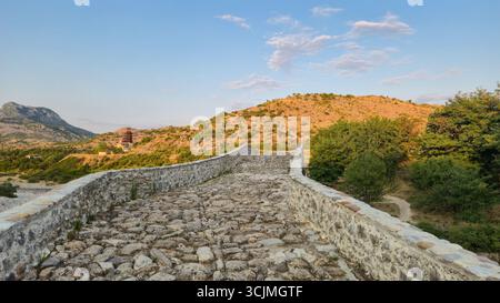Vue rapprochée du sentier de pierre sur le pont de Mesi pendant l'heure dorée, avec des collines en arrière-plan, près de Shkodër, Albanie. Banque D'Images