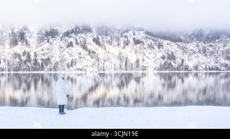 Une scène hivernale paisible et tranquille avec des montagnes enneigées se reflétant magnifiquement sur un lac immobile, Une petite femme dans une veste blanche regarde Banque D'Images