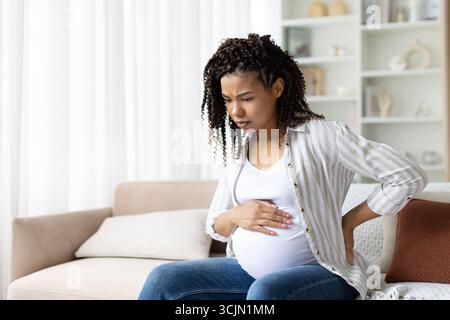 Femme noire enceinte assise sur le canapé avec la main sur le dos et le ventre Banque D'Images