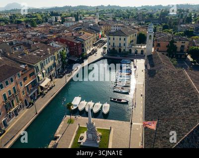 Vue aérienne des bateaux amarrés dans le port tranquille, encadrés par les bâtiments historiques et la tour de l'horloge de Lazise, Vénétie, Italie. Banque D'Images