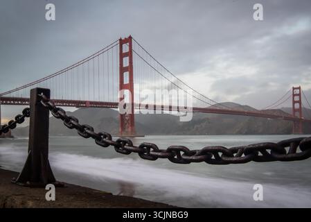 Vue sur l'emblématique Golden Gate Bridge rouge s'étend sur la baie sous un ciel nuageux, encadré par une chaîne industrielle lourde, San Francisco, Californie, États-Unis. Banque D'Images