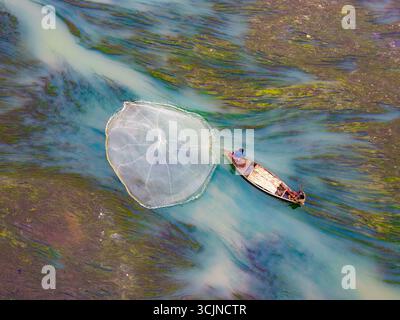Badalgachhi, Bangladesh - 09 décembre 2024 : vue aérienne d'un pêcheur solitaire jetant son filet depuis un bateau en bois sur la rivière, au milieu des eaux riches en algues. Banque D'Images
