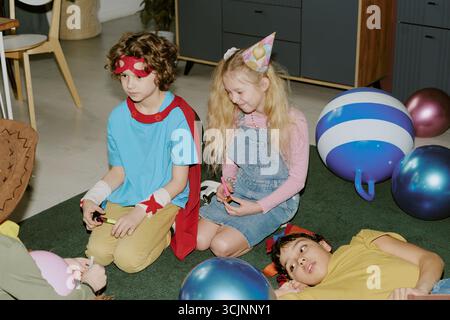Groupe d'enfants assis et couchés sur le sol pendant la fête d'anniversaire à l'intérieur Banque D'Images