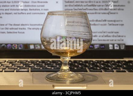 Londres, Royaume-Uni - 30 mai 2025 - verre de whisky ou snifter Brandy se tient sur ordinateur portable avec écran d'ordinateur portable en arrière-plan. Utilisez-le comme fond d'écran, Poster an Banque D'Images