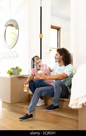 Jeune couple diversifié assis sur les escaliers à la maison, appréciant le café et la conversation Banque D'Images