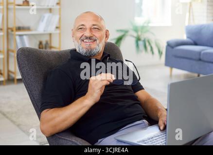 Homme senior heureux avec ordinateur portable à la chaise de bureau à la maison, prenant la pause avec des lunettes Banque D'Images
