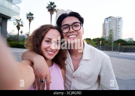 Couple multiracial utilisant le téléphone pour prendre un selfie regardant la caméra souriant debout dehors. Banque D'Images