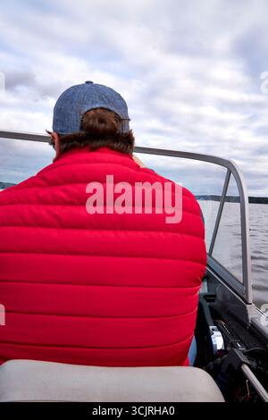 Homme dans une veste rouge et casquette bleue assis dans un bateau, profitant d'une balade paisible sur le lac sous un ciel nuageux. Concept de loisirs de plein air, voyage, et a Banque D'Images