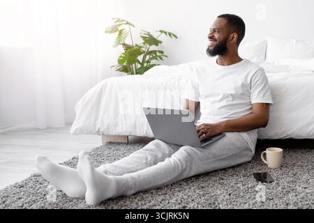 Joyeux jeune homme à barbe noire travaille sur un ordinateur portable, s'asseoir sur le sol dans la chambre à l'intérieur, regarder la fenêtre Banque D'Images