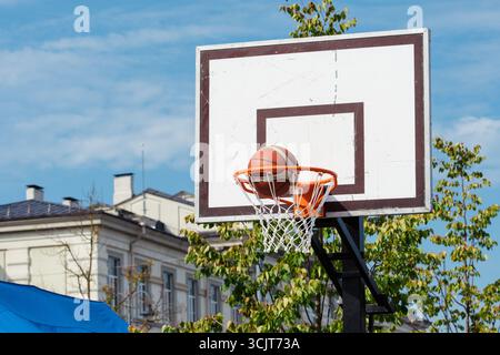 Panier de basket-ball extérieur avec une balle passant à travers le filet, placé contre un ciel bleu clair et des bâtiments urbains en arrière-plan Banque D'Images