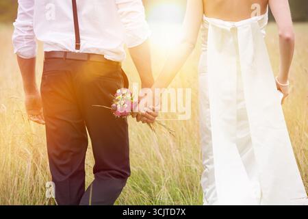 couple de jeunes mariés marchant main dans la main à travers de hautes herbes vers le soleil, vue arrière Banque D'Images