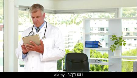 Médecin senior défilant portant une blouse de laboratoire visionnant des dossiers sur tablette dans la salle d'examen, avec stéthoscope Banque D'Images