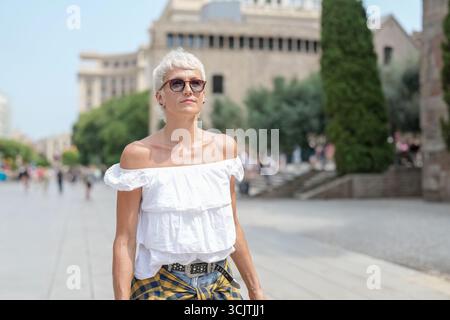 Une femme portant une chemise blanche et un pantalon jaune marche dans une rue. Elle porte des lunettes de soleil et profite de la journée ensoleillée Banque D'Images
