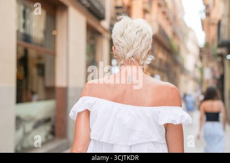 Une femme blonde portant une robe blanche marche dans une rue. Elle a un tatouage sur le dos Banque D'Images