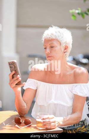Une femme est assise à une table avec un verre de bière et un téléphone portable. Elle regarde le téléphone tout en tenant le verre Banque D'Images