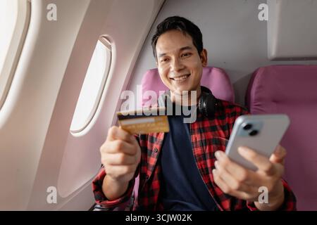 Jeune homme joyeux est assis sur un siège de fenêtre à l'intérieur d'un avion, tenant un smartphone dans une main et une carte de crédit dans l'autre. Il sourit, semblant le faire Banque D'Images
