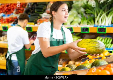 Jeune femme joyeuse ouvrière de supermarché dans le tablier tenant le melon mûr dans la section des fruits Banque D'Images