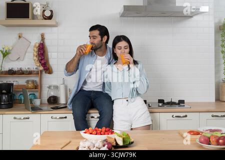 Jeune couple profitant de boire le matin ensemble dans la cuisine moderne, tenant des verres de jus d'orange frais avec ordinateur portable sur le comptoir, concept de vies saines Banque D'Images