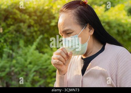 Vue de côté portrait d'une femme asiatique debout dans le parc et souffrant de toux avec protection de masque facial. Banque D'Images