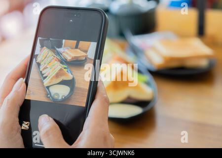 Gros plan des mains féminines à l'aide d'un téléphone intelligent prenant des photos de sandwichs et de café sur une table en bois. Mise au point sélective. Banque D'Images