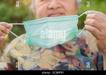 Vue rapprochée des mains de femme aînée mettant le masque facial contre le coronavirus sur le fond de la nature. Mise au point sélective. Banque D'Images