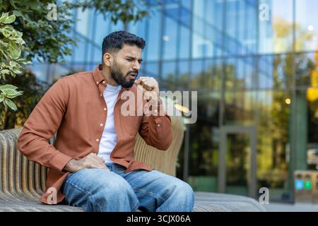 Homme barbu assis sur un banc dans la ville, toussant et couvrant sa bouche avec son poing, souffrant de maladie ou d'allergies pendant son trajet Banque D'Images