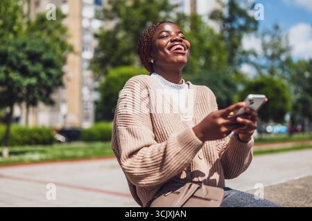 Jeune femme noire heureuse utilise un smartphone. Elle rit en envoyant des SMS au téléphone. Banque D'Images