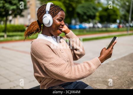 Portrait d'une femme afro-américaine joyeuse dans la vingtaine utilisant son téléphone portable à l'extérieur dans un cadre urbain, portant des écouteurs sans fil et souriant Banque D'Images
